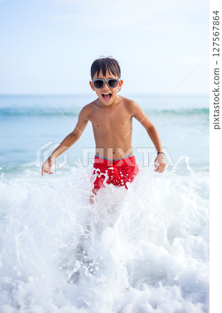 Boy Enjoying Splashing Water at Beach on Sunny Day Wearing Sunglasses and Red Swim Trunks 127567864