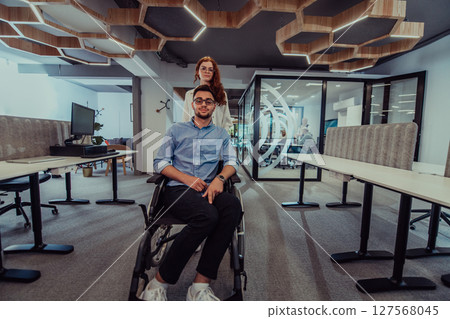 Young business colleagues, collaborative business colleagues, including a person in a wheelchair, walk past a modern glass office corridor, illustrating diversity, teamwork and empowerment in the Young business colleagues, collaborative business colleagues, including a person in a wheelchair, walk past a modern glass office corridor, illustrating diversity, teamwork and empowerment in the 127568045