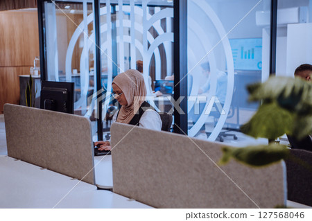In a modern office, a young Muslim entrepreneur wearing a hijab sits confidently and diligently works on her computer, embodying determination, creativity, and empowerment in the business world 127568046