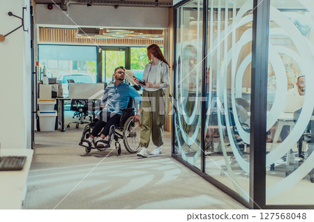 Young business colleagues, collaborative business colleagues, including a person in a wheelchair, walk past a modern glass office corridor, illustrating diversity, teamwork and empowerment in the Young business colleagues, collaborative business colleagues, including a person in a wheelchair, walk past a modern glass office corridor, illustrating diversity, teamwork and empowerment in the 127568048