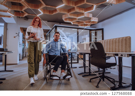 Young business colleagues, collaborative business colleagues, including a person in a wheelchair, walk past a modern glass office corridor, illustrating diversity, teamwork and empowerment in the 127568088