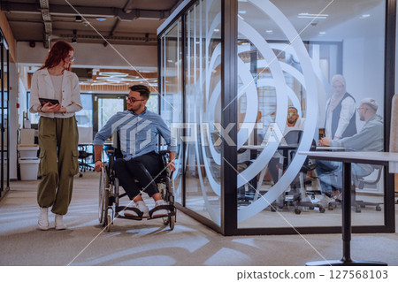 Young business colleagues, collaborative business colleagues, including a person in a wheelchair, walk past a modern glass office corridor, illustrating diversity, teamwork and empowerment in the 127568103