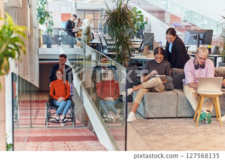 A company director assists his business colleague in a wheelchair, helping her navigate to their startup office, where they work alongside their diverse team of colleagues, emphasizing inclusivity and 127568135