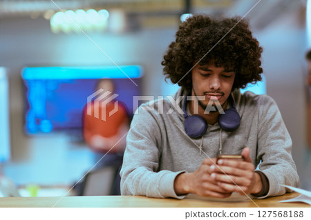 In a modern startup office, an African American man, balancing work and technology, utilizes headphones and a smartphone, emblematic of contemporary multitasking and productivity 127568188