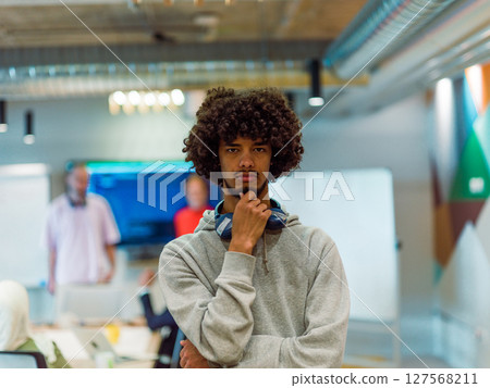 In a modern office environment, an African American young entrepreneur with headphones engages in work, while in the background, his dedicated colleagues exemplify teamwork and collaboration In a modern office environment, an African American young entrepreneur with headphones engages in work, while in the background, his dedicated colleagues exemplify teamwork and collaboration 127568211