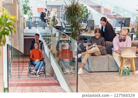 A company director assists his business colleague in a wheelchair, helping her navigate to their startup office, where they work alongside their diverse team of colleagues, emphasizing inclusivity and 127568329