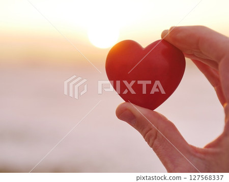 A hand holding a red heart in front of a sunset 127568337