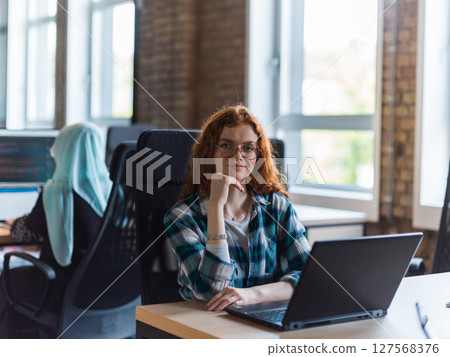 A young and successful businesswoman with vibrant orange hair engages in focused work within a modern office, showcasing her confidence and dynamic approach as she utilizes her laptop, emblematic of a 127568376