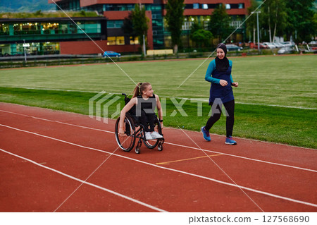 A Muslim woman in a burqa running together with a woman in a wheelchair on the marathon course, preparing for future competitions. A Muslim woman in a burqa running together with a woman in a wheelchair on the marathon course, preparing for future competitions. 127568690