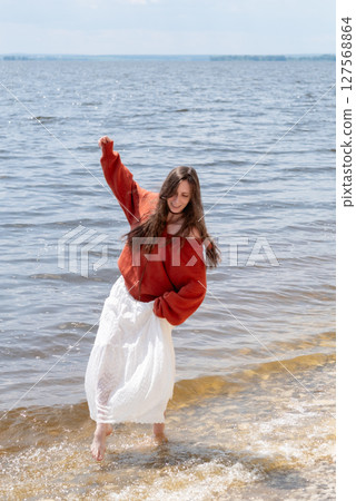 A young woman with long hair and blue eyes, in a white skirt and a terracotta sweater, runs barefoot on the water against the backdrop of the sea and laughs 127568864