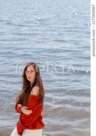 A young woman with long hair and blue eyes, in a white skirt and a terracotta sweater, stands barefoot in the water against the backdrop of the sea 127568867