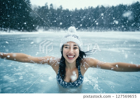 Smiling woman in bikini and winter hat enjoying ice swimming in a frozen lake. Water splashes around her in a joyful moment surrounded by snowy landscape and forest in the background. Banner 127569655