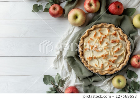 Delicious homemade apple pie with golden crust surrounded by fresh apples and green leaves on rustic white wooden background. Perfect autumn dessert or bakery product presentation. Copy space. Banner 127569658