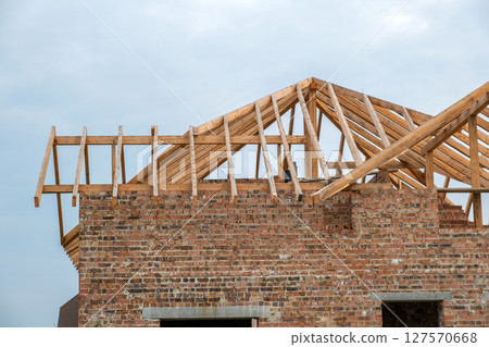 Building site with unfinished brick house with wooden roofing frame for future roof under construction. Building site with unfinished brick house with wooden roofing frame for future roof under construction. 127570668