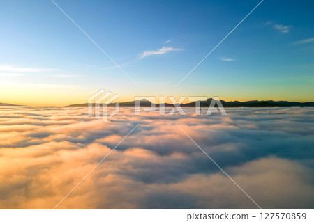 Aerial view of yellow sunset over white puffy clouds with distant mountains on horizon. Aerial view of yellow sunset over white puffy clouds with distant mountains on horizon. 127570859