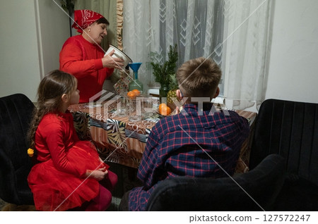 A grandmother pours juice for her grandchildren at a table set for a small gathering. Warmth and family connection are evident. A grandmother pours juice for her grandchildren at a table set for a small gathering. Warmth and family connection are evident. 127572247