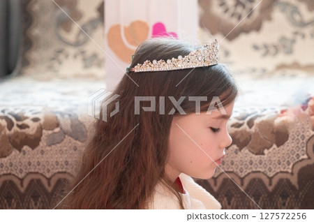 A young girl with long brown hair wears a sparkly tiara, appearing pensive and serene. A young girl with long brown hair wears a sparkly tiara, appearing pensive and serene. 127572256