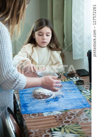 A mother and daughter bake together, sifting flour onto a dough. A moment of shared learning and family bonding in the kitchen. 127572261