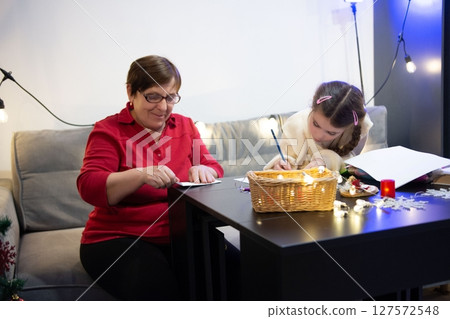 Grandmother and granddaughter spend quality time together writing letters and drawing, surrounded by cozy holiday lights. 127572548