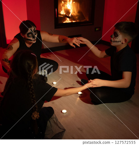 A Halloween night seance. Three figures, faces painted, hold hands around flickering candlelight, creating an eerie atmosphere. 127572555