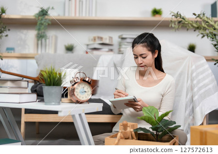 Moving and Organizing. Young woman writing notes while preparing for relocation in a cozy living space. Moving and Organizing. Young woman writing notes while preparing for relocation in a cozy living space. 127572613