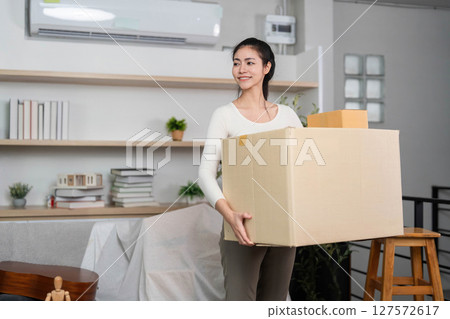 Moving and Strength. Young woman carrying a large box while relocating in her stylish home. 127572617