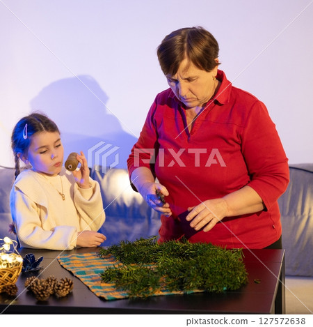 Grandmother and granddaughter crafting a festive evergreen arrangement together, sharing a moment of intergenerational bonding and creativity. 127572638