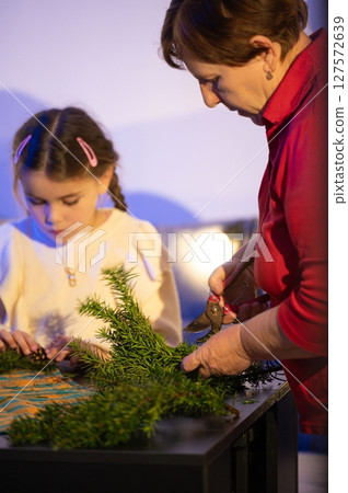 A woman and child work together, crafting a festive arrangement using greenery and small clippers. A woman and child work together, crafting a festive arrangement using greenery and small clippers. 127572639