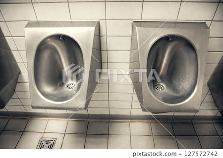 Stainless steel urinals in a public restroom with white tiled walls. The industrial and functional 127572742