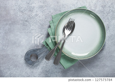 an empty plate and cutlery laid out, on a cutting board, with a green napkin, a gray table, top view, 127573784