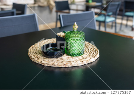 Green glass jar and sugar packets on woven placemat. Cafe table setting, casual dining ambiance and stylish hospitality decor. Green glass jar and sugar packets on woven placemat. Cafe table setting, casual dining ambiance and stylish hospitality decor. 127575397