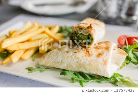 Fresh tortilla wrap served with golden fries and arugula on a white plate close-up. Casual dining, fast lunch and tasty street food in a modern restaurant setting. Fresh tortilla wrap served with golden fries and arugula on a white plate close-up. Casual dining, fast lunch and tasty street food in a modern restaurant setting. 127575422