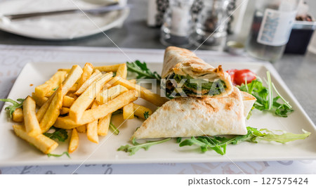 Fresh tortilla wrap served with golden fries and arugula on a white plate close-up. Casual dining, fast lunch and tasty street food in a modern restaurant setting. 127575424
