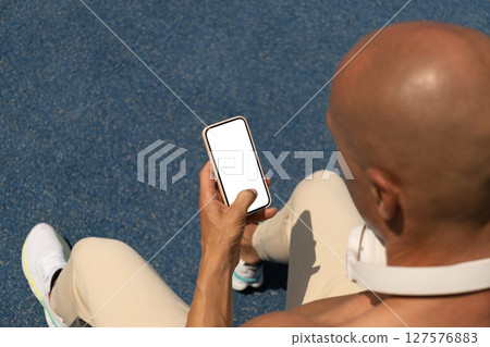 Sport man sitting and using smartphone with white blank screen. Checks apps with sport fitness progress and healthy. Over shoulder view. Space for text. Sport man sitting and using smartphone with white blank screen. Checks apps with sport fitness progress and healthy. Over shoulder view. Space for text. 127576883