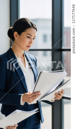 A woman in suit reading documents while standing near window. A woman in suit reading documents while standing near window. 127577646