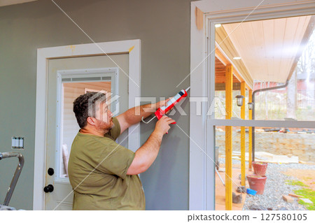 Carpenter carefully seals gaps around windows with caulk while renovating room in house. 127580105