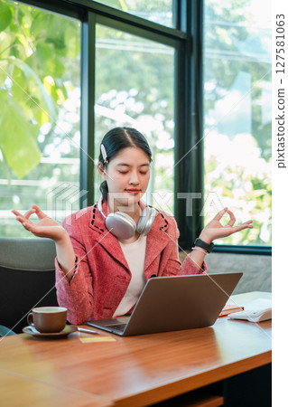 A young woman meditates with headphones at a laptop in a modern office, surrounded by natural light and greenery. 127581063