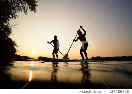 Paddleboarders Gliding Across Calm Water at Sunset With Orange and Purple Skies Paddleboarders Gliding Across Calm Water at Sunset With Orange and Purple Skies 127586806