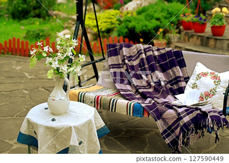 Apple blossoms on a table in front of a sofa swing in the courtyard of a country house. Apple blossoms on a table in front of a sofa swing in the courtyard of a country house. 127590449