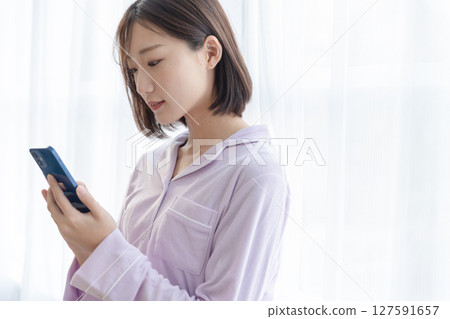 A young woman wearing pajamas sitting by the window and using a smartphone 127591657