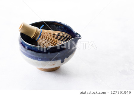 Japanese tea bowl glazed with blue and white color and Chasen or Bamboo whisk, an equipment for preparing matcha green tea on white wooden table. Japanese tea bowl glazed with blue and white color and Chasen or Bamboo whisk, an equipment for preparing matcha green tea on white wooden table. 127591949