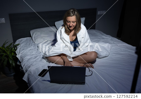 Woman Working on Laptop in Bed While Wrapped in a Blanket in a Cozy Bedroom at Night 127592214