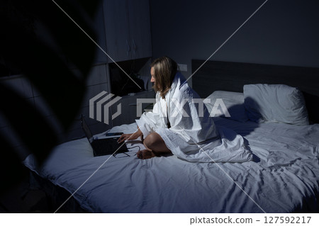 Person Working on a Laptop in Bed Under a Blanket During Nighttime Hours 127592217