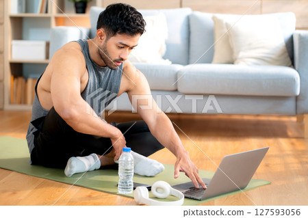 Tired young asian man sitting on a yoga mat after a home workout wiping sweat in living room. 127593056
