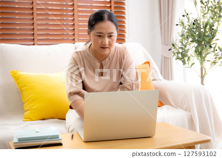 Asian woman working or studying online using a laptop sitting on sofa in living room at home. 127593102