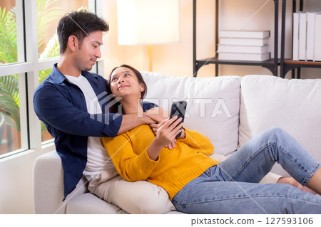 Happy young asian couple relax together in living room at home on cozy sofa while using smartphone. 127593106