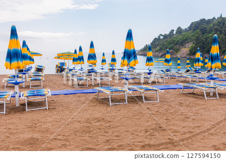 Tranquil morning at a beach with blue umbrellas and loungers. Coastal vacation, summer relaxation and peaceful seaside tourism. Tranquil morning at a beach with blue umbrellas and loungers. Coastal vacation, summer relaxation and peaceful seaside tourism. 127594150