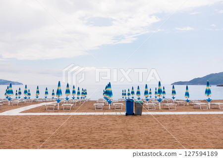 Empty beach with rows of closed umbrellas and sun loungers on a cloudy day. Coastal relaxation, off-season atmosphere and summer tourism infrastructure. Empty beach with rows of closed umbrellas and sun loungers on a cloudy day. Coastal relaxation, off-season atmosphere and summer tourism infrastructure. 127594189
