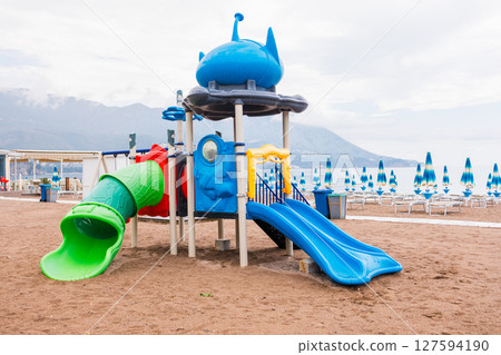Colorful playground by the Adriatic Sea with beach umbrellas and mountain view. Summer vacation, seaside recreation and family tourism in Montenegro. Colorful playground by the Adriatic Sea with beach umbrellas and mountain view. Summer vacation, seaside recreation and family tourism in Montenegro. 127594190