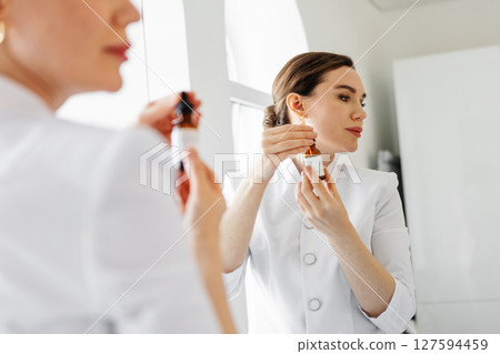Woman in white coat holding anti-aging serum and dropper near face, in front of mirror in beauty clinic, demonstrating professional skincare product for facial treatment and healthy glowing skin Woman in white coat holding anti-aging serum and dropper near face, in front of mirror in beauty clinic, demonstrating professional skincare product for facial treatment and healthy glowing skin 127594459